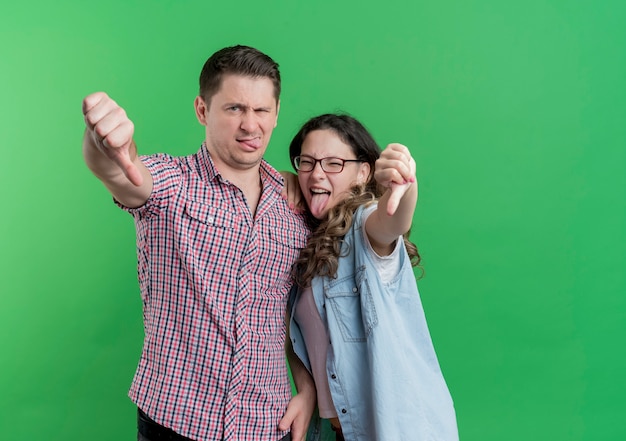 Young couple man and woman in casual clothes  displeased showing thumbs down standing over green wall