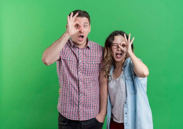 Young couple man and woman in casual clothes  making ok signs looking through this signs standing over green wall