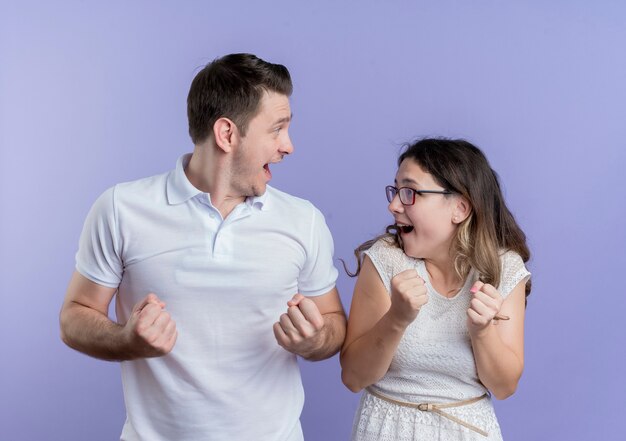 Young couple man and woman clenching fists happy and excited standing over blue wall