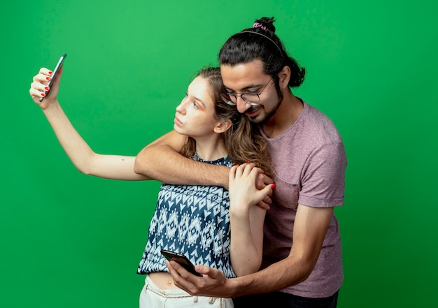 Free photo young couple man and woman, happy man hugging his girlfriend while she taking picture of them using smartphone standing over green background