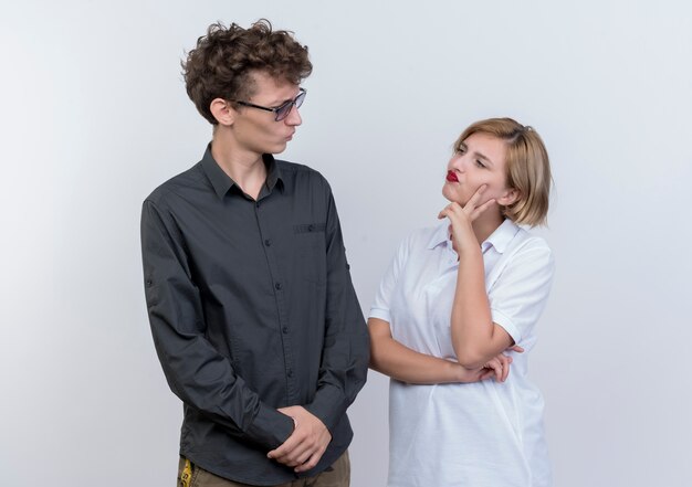 Young couple man and woman looking at each other with skeptic expression standing over white wall