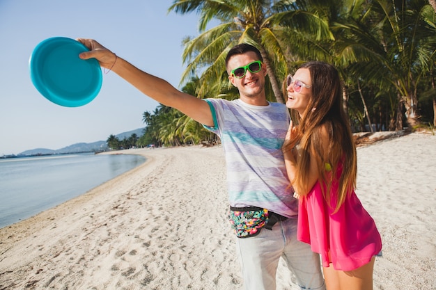 Young couple man and woman playing flying disk on tropical beach, summer vacation, love, romance, happy mood, smiling, having fun, hipster outfit, sunglasses, denim shorts, sunny, positive mood