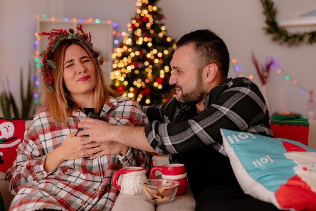 young couple man and woman with smartphone sitting on a couch with cups of tea quarreling decorated room with christmas tree in the background