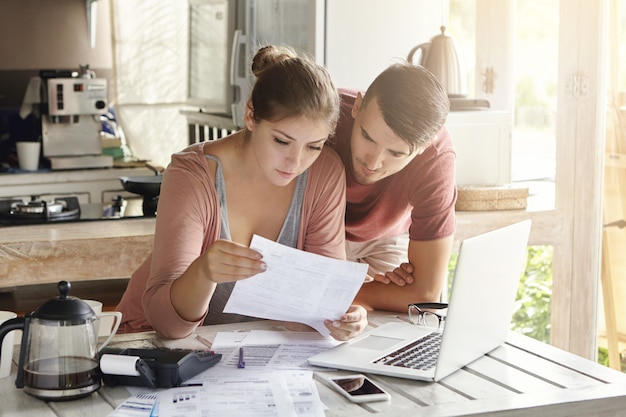 Free photo young couple managing finances, reviewing their bank accounts using laptop computer and calculator at modern kitchen. woman and man doing paperwork together