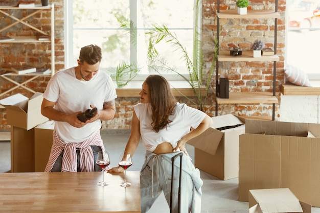 Free photo young couple moved to a new house or apartment. drinking red wine, smiling and relaxing after cleaning and unpacking. look happy and confident. family, moving, relations, first home concept.