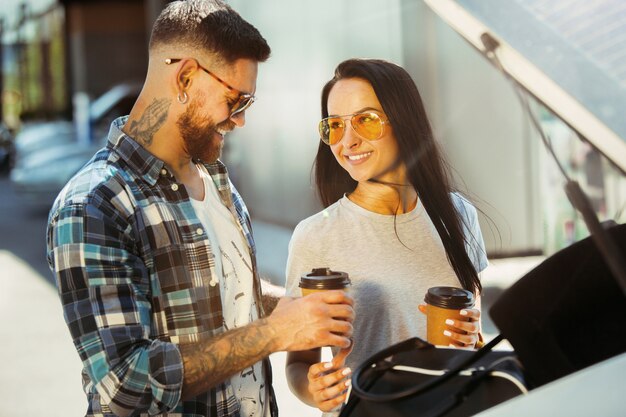 Young couple preparing for vacation trip on the car in sunny day. Woman and man drinking coffee and ready for going to sea or ocean.