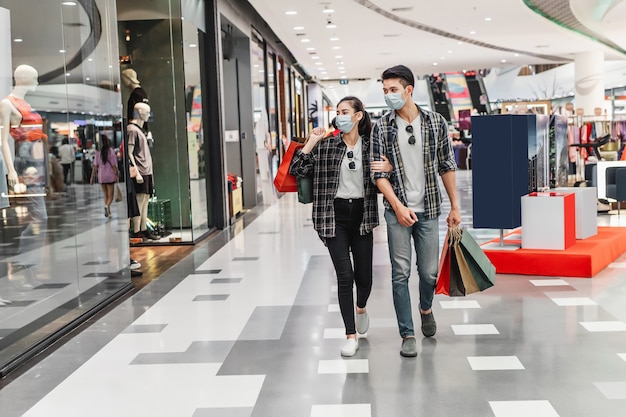 Young couple in protection mask holding multiple paper shopping bag walking in the corridor of large shopping mall