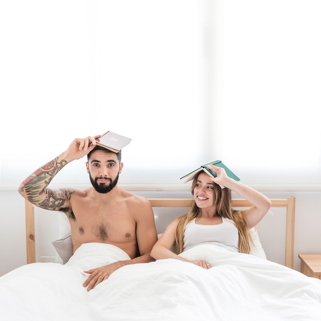 Free photo young couple sitting on bed holding book over their head