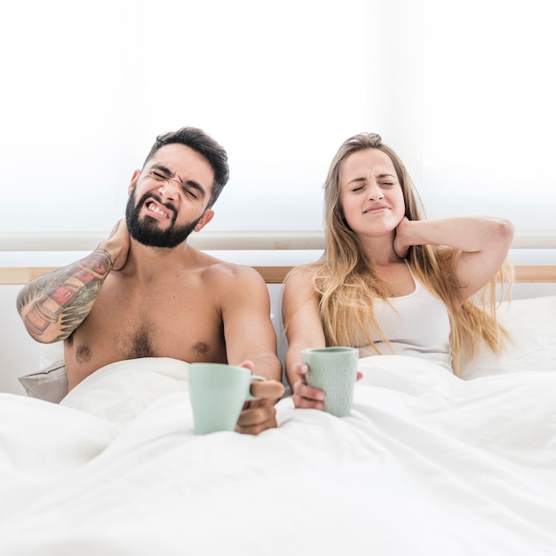 Free photo young couple sitting on bed with cup of coffee suffering from neck pain