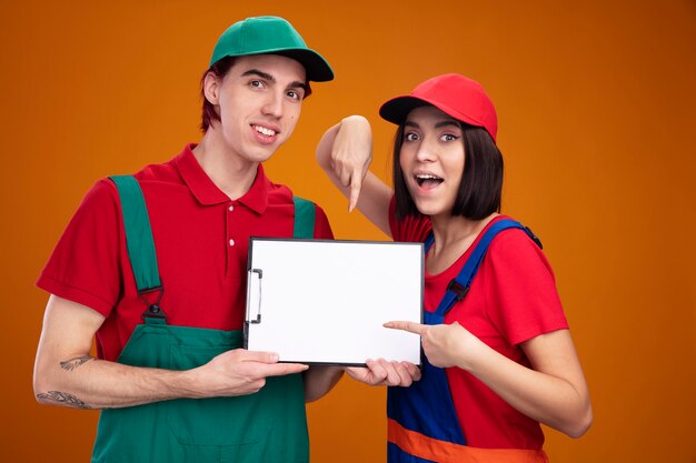 Young couple smiling guy excited girl in construction worker uniform and cap guy showing clipboard girl pointing at it both looking at camera isolated on orange wall