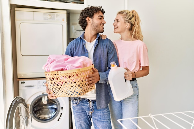 Young couple smiling happy holding laundry basket and detergent bottle at home.