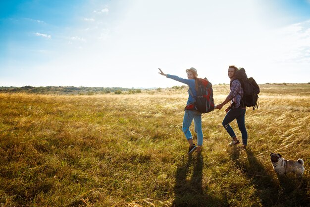 Young couple of travelers walking in field with pug dog