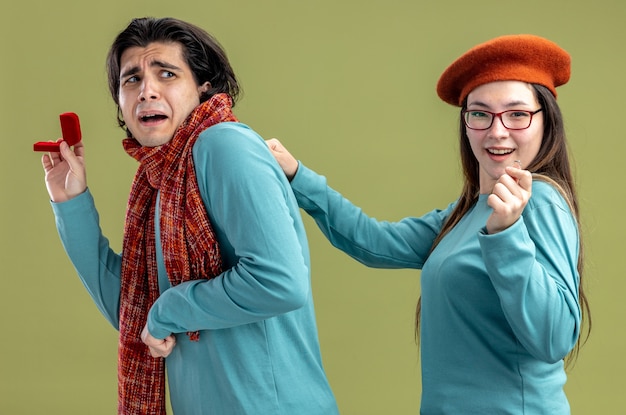 Free Photo young couple on valentines day guy wearing scarf girl wearing hat scared guy giving wedding ring to pleased girl isolated on olive green background