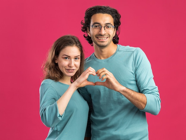 Young couple wearing pajamas smiling man wearing glasses pleased woman both looking at front doing heart sign together isolated on pink wall