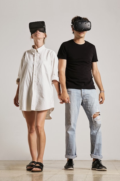 Free photo a young couple wearing vr headsets holding hands looking in different directions in a room with white walls and wooden floors