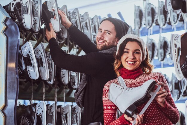 Young couple wearing warm clothes standing near rack with many pairs of skates, choosing his size