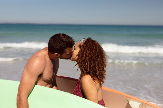 Young couple with surfboard kissing each other on beach in the sunshine