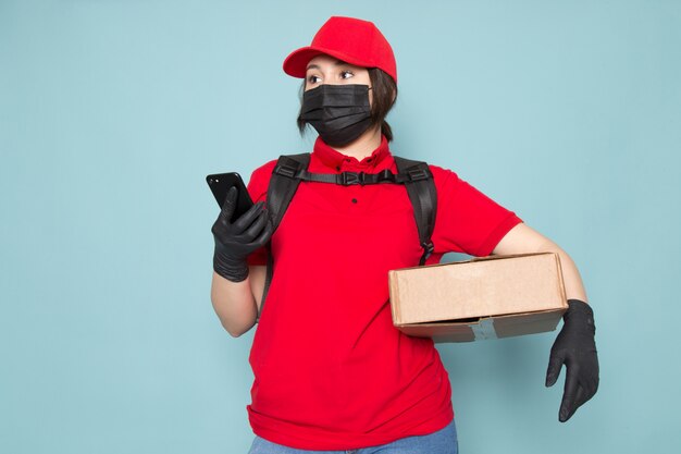 young courier in red polo red cap black sterile protective mask black backpack holding package using phone on blue