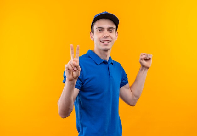 Young courier wearing blue uniform and blue cap doing happy peace and fist