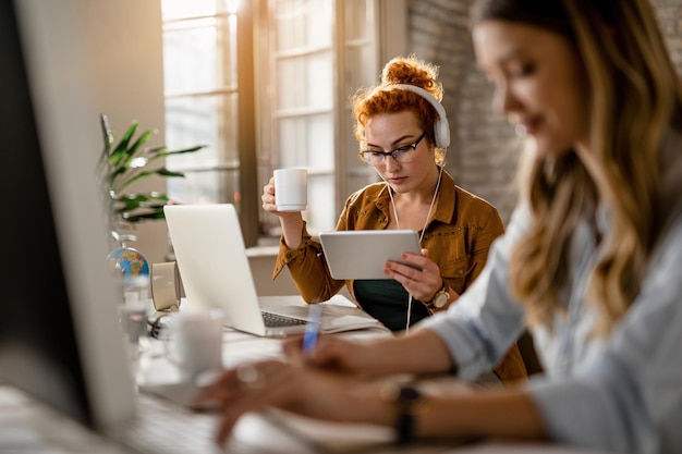 Free Photo young creative businesswoman wearing headphones while surfing the net on digital tablet and drinking coffee in the office