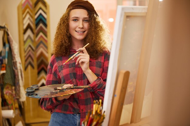 Young curly artist looks happy and holds a palette with oil paints