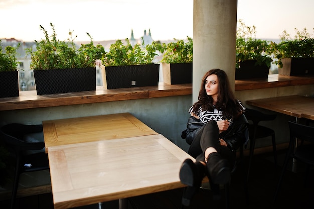 Free photo young curly woman in leather jacket in a bar put her legs on table