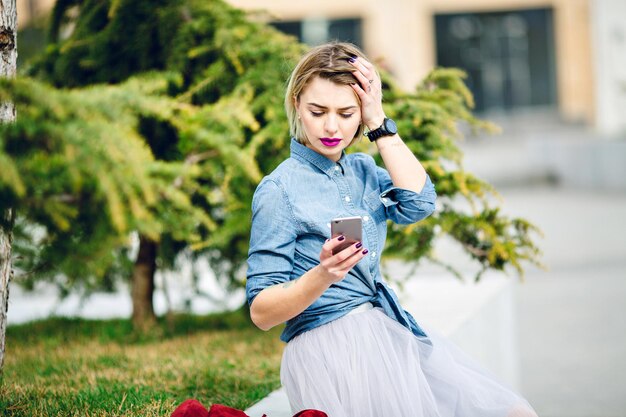 Young cute blond girl with short hair and bright pink lips sitting in a park and reading a message on her smartphone wearing denim blue shirt and grey tulle skirt.