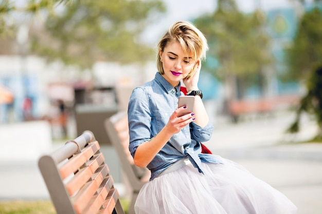 Young cute blond girl with short hair sitting on a wooden bench and looking at smartphone wearing denim blue shirt and grey tulle skirt combing her hair behind ear.