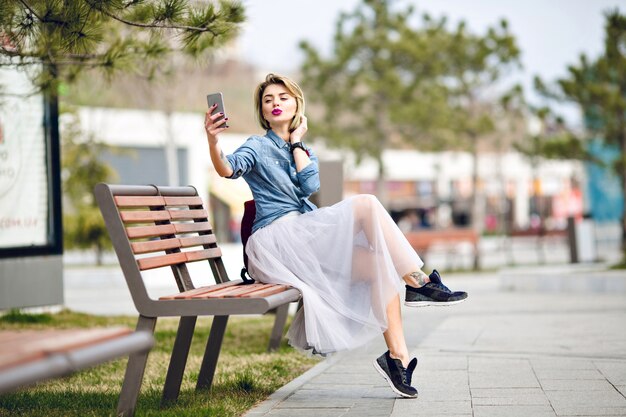 Young cute blond woman with short hair and bright pink lips sitting on wooden bench making a selfie with kissing lips