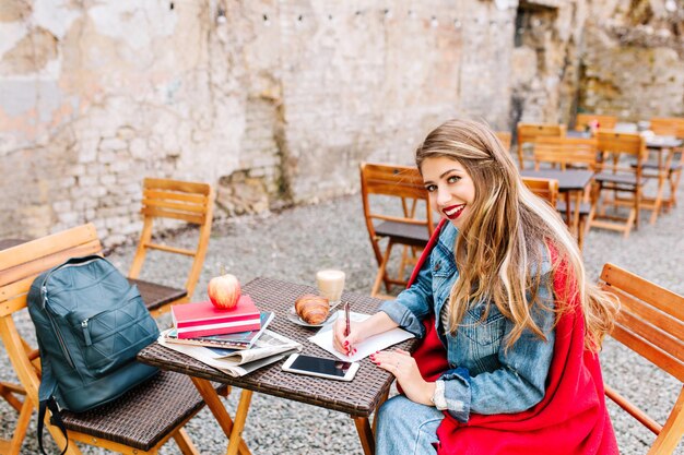 Free photo young cute fair-haired girl working on the veranda during lunch. pretty student makes notes sitting in a open-air cafe near the brick building. denim clothes, red lipstick, busy morning