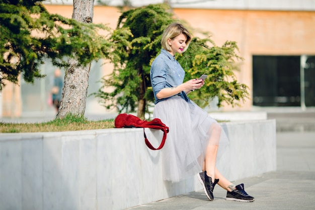 Young cute smiling blond woman with short hair and bright pink lips sitting in a park and reading a message on her smartphone