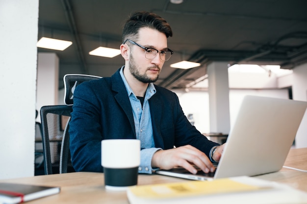 Young  dark-haired man is working at the table in office. He wears blue shirt wi8th black jacket. He is typing on laptop seriously.