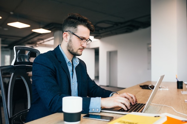 Free photo young dark-haired man is working at the table in office. he wears blue shirt with black jacket. he is typing on laptop and looks busy.