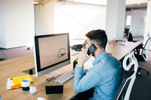 Young dark-haired man is working with a computer and talking on the phone at his desktop in office. He wears blue shirt and looks busy.