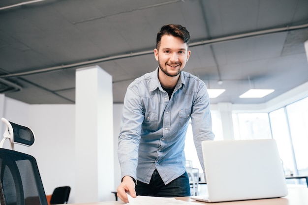 Young dark-haired man is working with a laptop in office. He wears blue shirt. He is smiling to the camera.