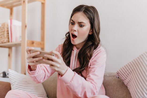 Young dark-haired model sits in her room and takes selfie with surprised facial expression