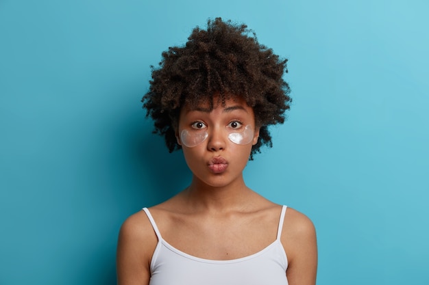 Free photo young dark skinned woman with afro hair applies hydrogel silver patches under eyes, reduces puffiness, removes dark circles. skin care concept