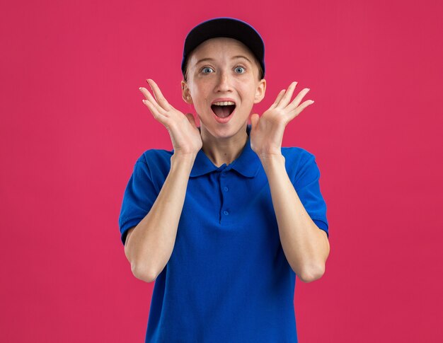 Young delivery girl in blue uniform and cap  happy and surprised with arms raised standing over pink wall