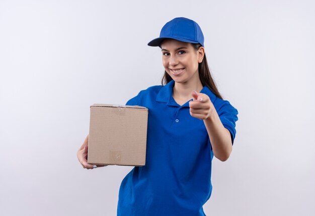 Young delivery girl in blue uniform and cap holding box package smiling confident pointing with finger to camera 