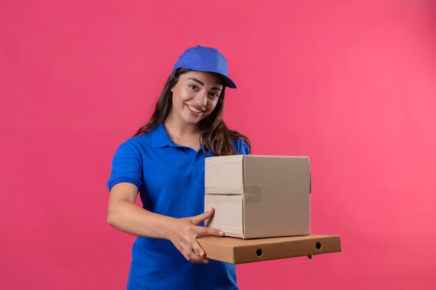 Young delivery girl in blue uniform and cap holding cardboard boxes looking at camera smiling confident happy and positive standing over pink background