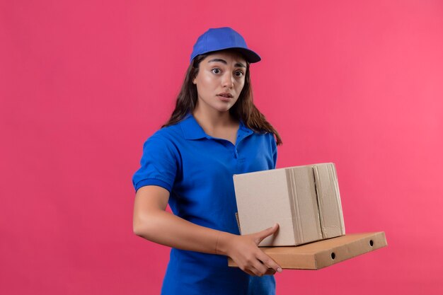 Young delivery girl in blue uniform and cap holding cardboard boxes looking surprised and amazed standing over pink background