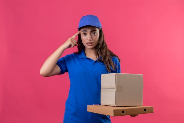 Young delivery girl in blue uniform and cap holding cardboard boxes pointing temple for mistake looking anxious standing over pink background