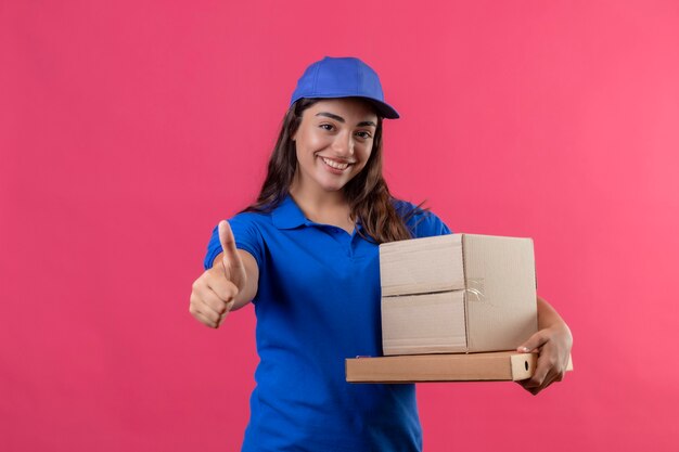 Young delivery girl in blue uniform and cap holding cardboard boxes smiling cheerfully showing thumbs up standing over pink background