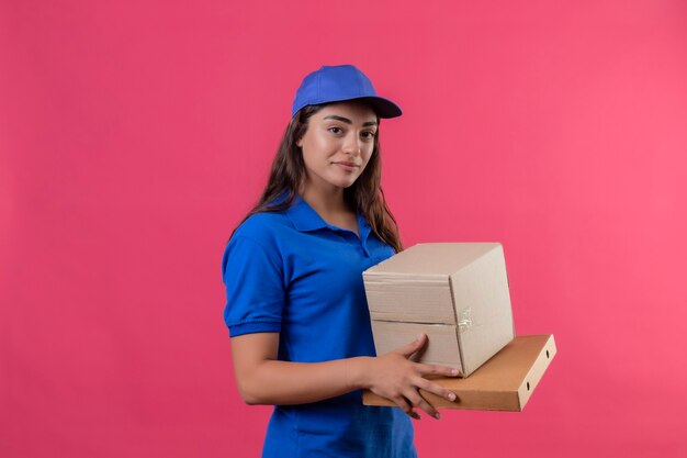 Young delivery girl in blue uniform and cap holding cardboard boxes smiling confident positive and happy standing over pink background