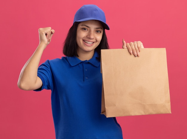 Young delivery girl in blue uniform and cap holding paper package clenching fist happy and excited standing over pink wall