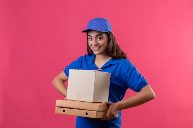 Young delivery girl in blue uniform and cap holding pizza boxes and box package looking at camera smiling cheerfully happy and positive standing over pink background