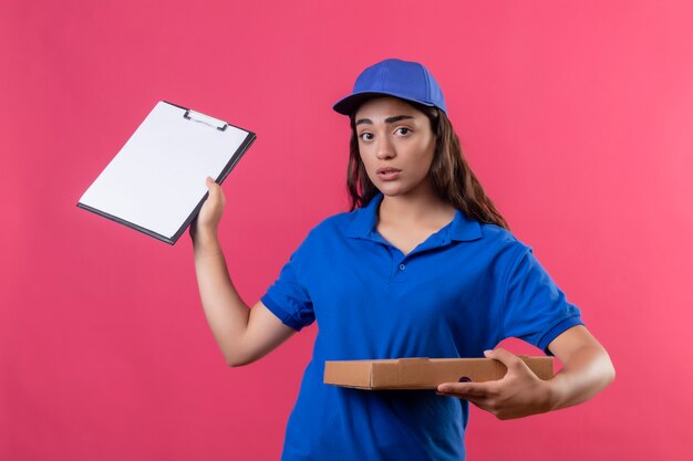 Young delivery girl in blue uniform and cap holding pizza boxes showing clipboard looking anxious standing over pink background