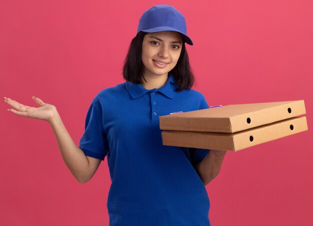 Young delivery girl in blue uniform and cap holding pizza boxes  smiling friendly with raised arm standing over pink wall