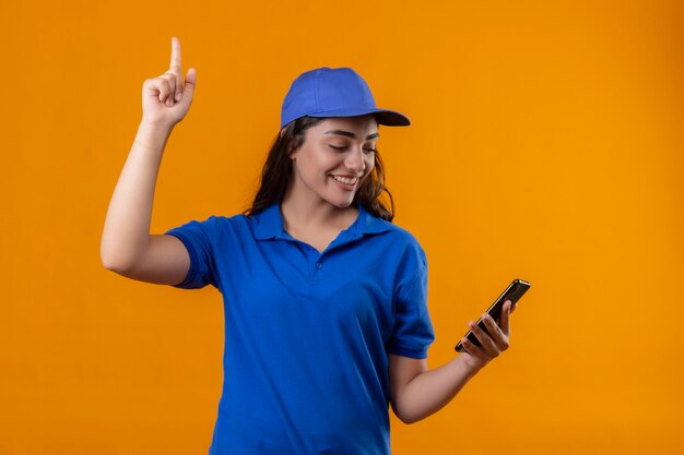 Young delivery girl in blue uniform and cap holding smartphone in hand looking at it pointing finger up having great idea smiling confident standing over yellow background