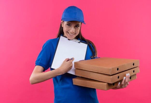 Young delivery girl in blue uniform and cap holding stack of pizza boxes and clipboard with blank pages asking for signature 